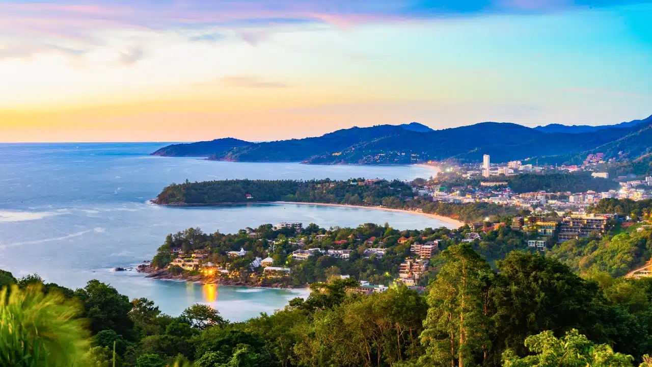 Panoramic view of Phuket, Thailand, featuring tropical beaches, lush green hills, and turquoise waters along the Andaman Sea at sunset.