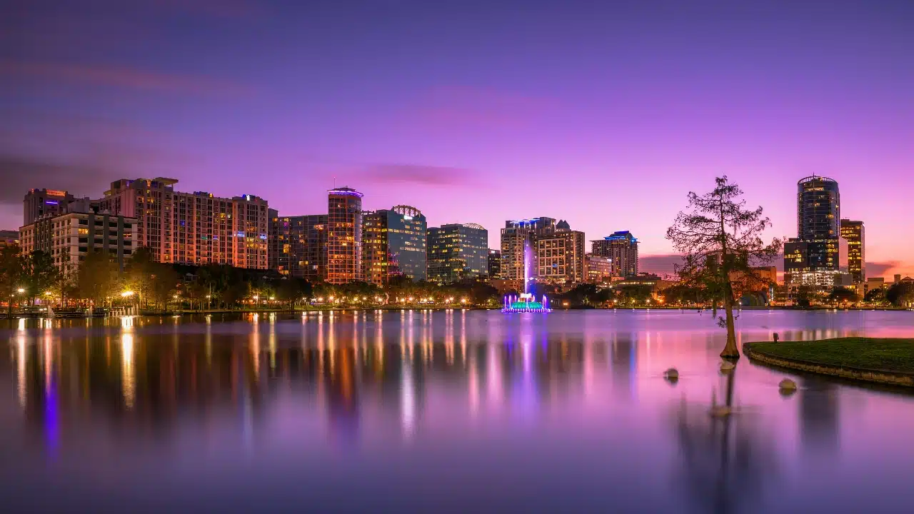 Orlando skyline at sunset viewed across Lake Eola, with colorful city lights reflecting on the water and the illuminated fountain in downtown Orlando, Florida.