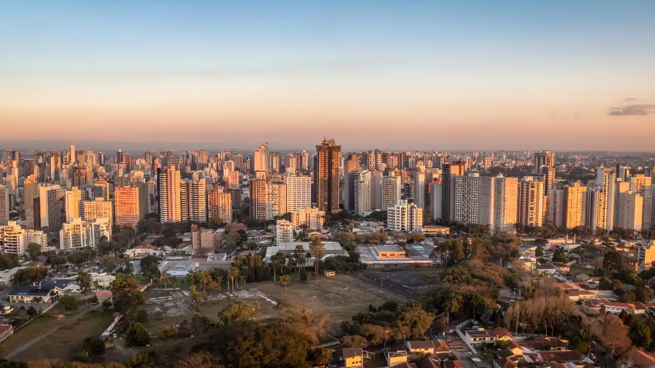 Panoramic view of Curitiba, Brazil, showing the city's dense skyline, residential neighborhoods, and urban landscape at sunset.