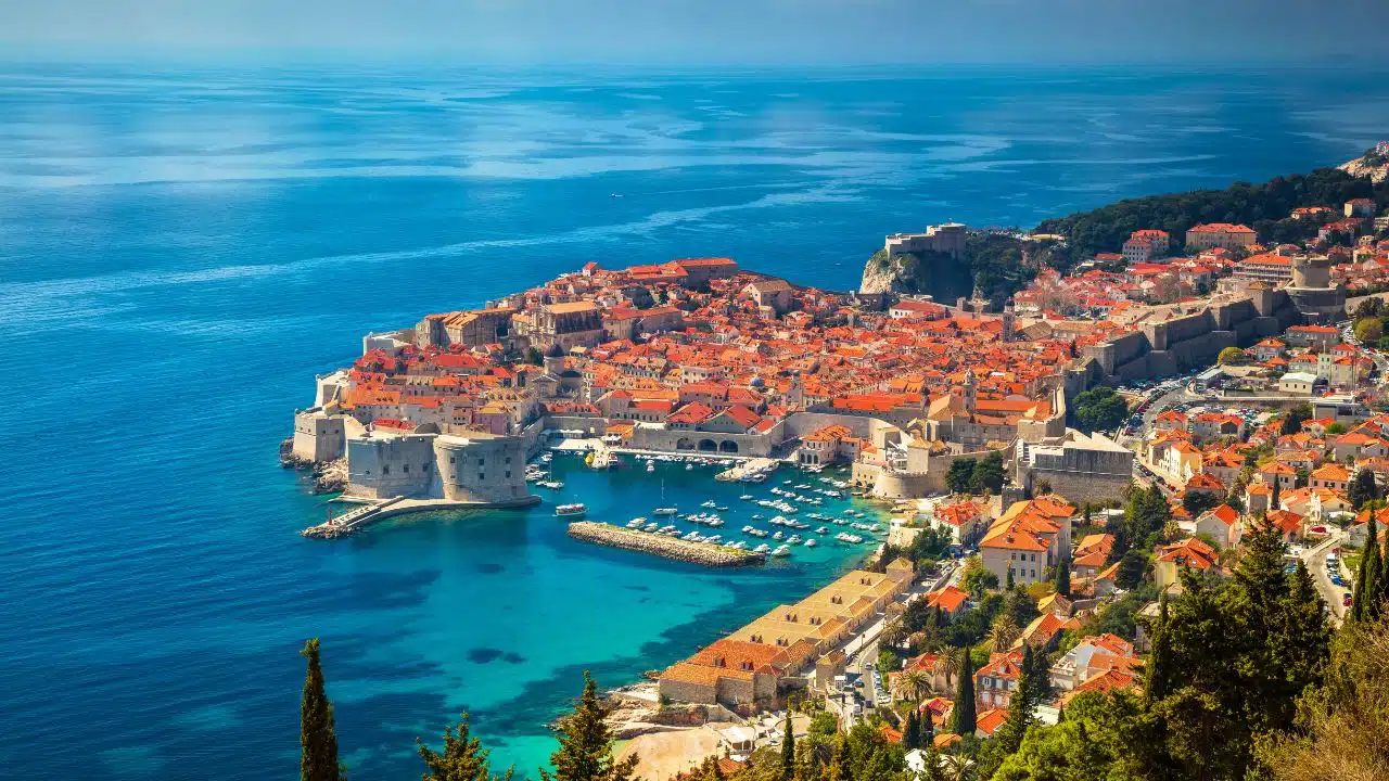 Aerial view of Dubrovnik, Croatia, featuring the historic Old Town, medieval city walls, terracotta rooftops, turquoise Adriatic Sea, and harbor.